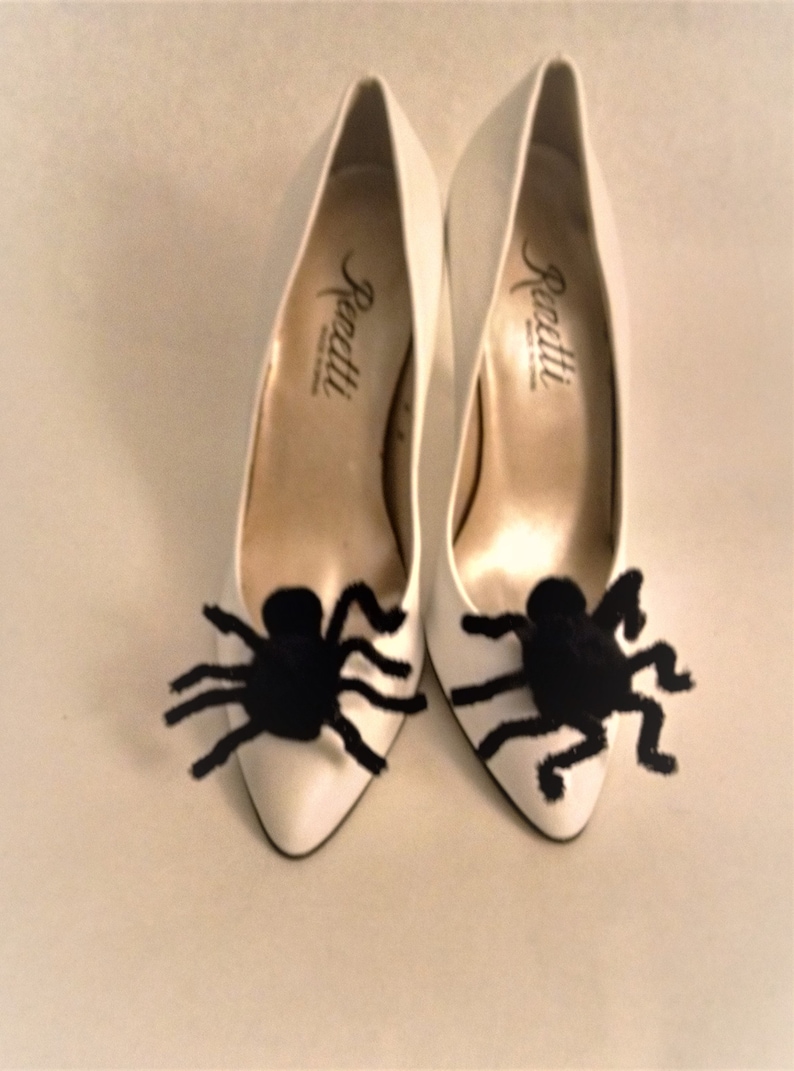 Pode incluir: Um par de sapatos brancos de salto alto com enfeites de aranha peluda preta nos dedos. Os sapatos s&atilde;o etiquetados como "Renatti".