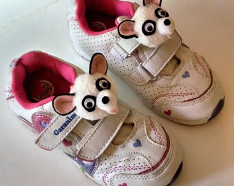 May include: A pair of white children's sneakers with pink accents and a fuzzy panda charm on each shoe. The shoes have a velcro strap and a heart design.