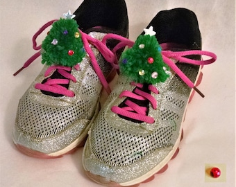 May include: A pair of silver glitter sneakers with pink laces and black collars. Each shoe has a small green Christmas tree ornament on the laces, decorated with colorful beads and a white star. A red ladybug-shaped embellishment is also visible.