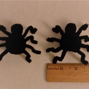 Pode incluir: Duas decora&ccedil;&otilde;es de aranha de pel&uacute;cia preta com oito patas cada. As aranhas medem aproximadamente 7,5 cm de largura e est&atilde;o sobre uma superf&iacute;cie branca ao lado de uma r&eacute;gua de madeira.