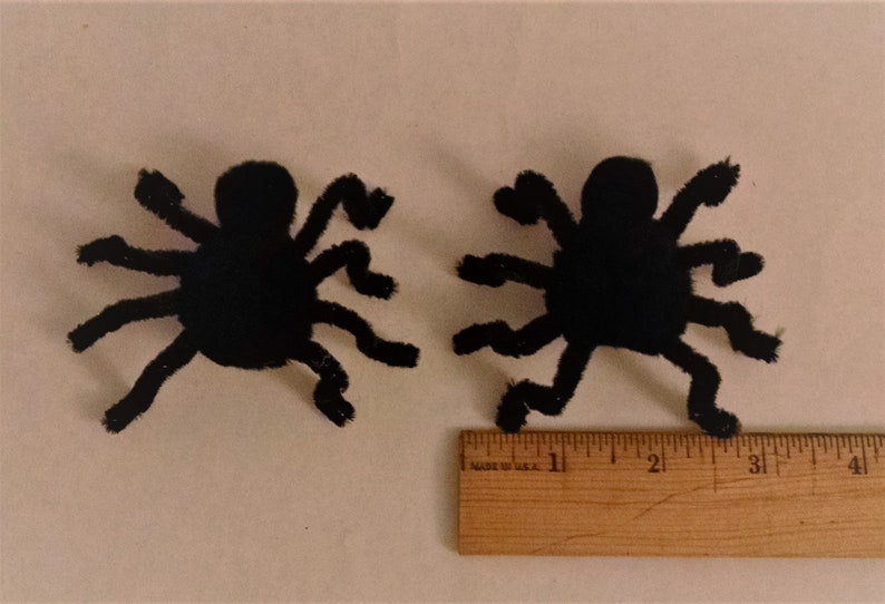 Pode incluir: Duas decora&ccedil;&otilde;es de aranha de pel&uacute;cia preta com oito patas cada. As aranhas medem aproximadamente 7,5 cm de largura e est&atilde;o sobre uma superf&iacute;cie branca ao lado de uma r&eacute;gua de madeira.