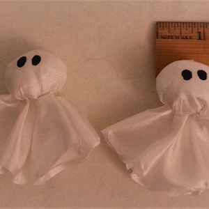 Pode incluir: Dois enfeites de fantasmas de tecido branco com olhos pretos. Os fantasmas s&atilde;o feitos de um tecido transparente e t&ecirc;m uma cabe&ccedil;a redonda.