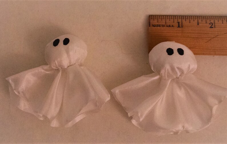 Pode incluir: Dois enfeites de fantasmas de tecido branco com olhos pretos. Os fantasmas s&atilde;o feitos de um tecido transparente e t&ecirc;m uma cabe&ccedil;a redonda.