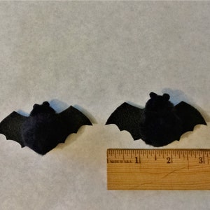 Pode incluir: Dois morcegos de feltro preto com textura felpuda, com aproximadamente 5 cm de comprimento, s&atilde;o exibidos em uma superf&iacute;cie branca com uma r&eacute;gua de madeira para escala.