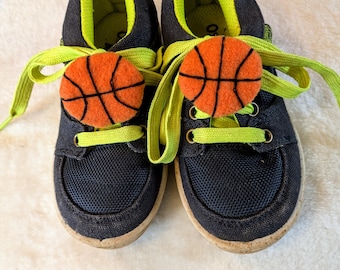 May include: A pair of navy blue children's sneakers with bright green laces and inner lining. Each shoe has a felt basketball applique on the laces. The soles are a light brown color.
