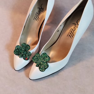 May include: A pair of white high-heeled shoes with green glitter shamrock embellishments. The shoes are made by the brand "Renati" and are made in Spain.