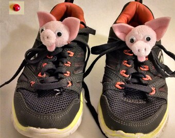 May include: A pair of black and green sneakers with pink pig shoelace charms. The pig charms have googly eyes and tongues sticking out.