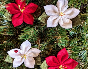 May include: Four fabric flower ornaments featuring five-petaled blossoms in red and white velvet-like material. Each flower has a central cluster of yellow stamens and is attached to a pair of dark green velvet leaves. One white flower has a red bead.