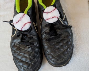 以下が含まれることがあります： 野球をテーマにした靴ひも飾り付きの黒と金のスポーツシューズのペア。靴はつま先にキルト模様があり、内側はネオングリーンです。各靴には、小さな白い野球が靴ひもに付いています。