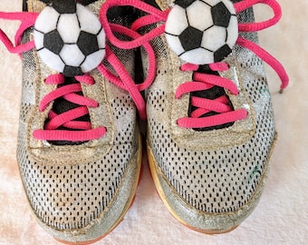 May include: A pair of silver and black athletic shoes with pink laces. Each shoe has a soccer ball decoration on the laces. The shoes have a mesh design and a red sole.