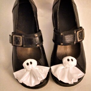 Pode incluir: Um par de sapatos de couro preto com decora&ccedil;&otilde;es de fantasmas brancos nos dedos. Os sapatos t&ecirc;m uma fivela e uma ponta pontiaguda.