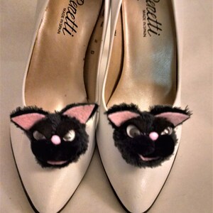 Pode incluir: Um par de sapatos brancos de salto alto com carinhas de gatinhos pretos e peludos presas aos dedos. Os sapatos s&atilde;o etiquetados com "Renetti Made in Spain."