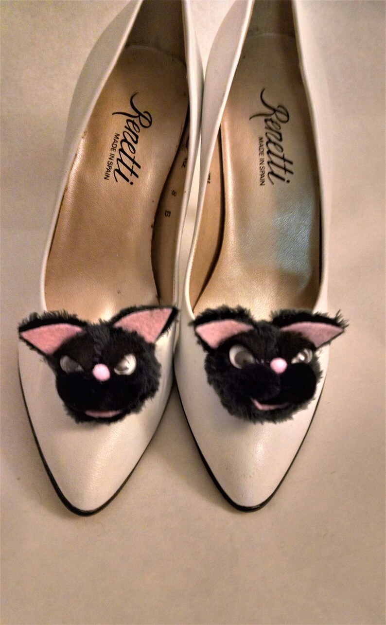 Pode incluir: Um par de sapatos brancos de salto alto com carinhas de gatinhos pretos e peludos presas aos dedos. Os sapatos s&atilde;o etiquetados com "Renetti Made in Spain."