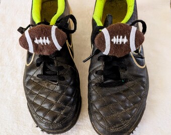 May include: Pair of black soccer cleats with a quilted design, black laces, and a gold swoosh. Each shoe has a brown and white football-shaped accessory attached to the laces. The interior of the shoes is lime green.