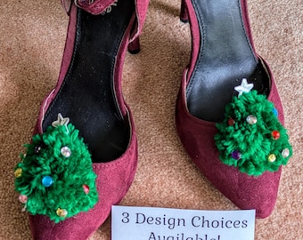 May include: A pair of burgundy-colored high-heeled shoes with a pointed toe. Each shoe has a small, green, pom-pom Christmas tree attached to the toe. The trees are decorated with colorful gems and a star. The text "3 Design Choices Available!" is on a white card.