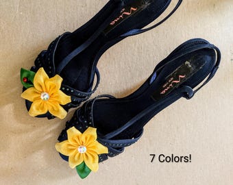 May include: A pair of dark navy blue Nina New York slingback heels featuring thin straps adorned with small dark crystals and a prominent yellow fabric flower accent on the toe. The insole displays the Nina brand logo and the text 7 Colors!