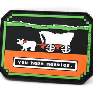 May include: A black, green, and orange patch with pixelated graphics. The design features a white ox pulling a covered wagon, with the text "you have measles." The patch has a green border and the text "STINKINGPATCHES.COM".