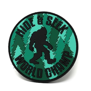 May include: A green and black circular patch featuring a silhouette of Bigfoot walking through a forest. The text "Hide & Seek World Champ" is written around the edge of the patch.