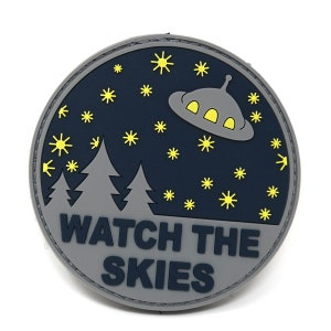 May include: A round, grey patch with a dark blue background. The patch features a cartoon UFO flying over a forest with stars in the sky. The text "WATCH THE SKIES" is printed in white on the bottom of the patch.