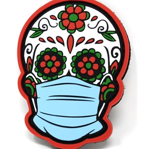 May include: A white sugar skull with red and green floral designs, wearing a light blue face mask. The skull is outlined in red.