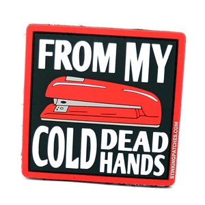 Op de afbeelding: Een vierkante patch in rood en zwart met witte tekst die "From my cold dead hands" luidt en een afbeelding van een rode nietmachine. De tekst "Stinkingpatches.com" staat in de rechterbenedenhoek.