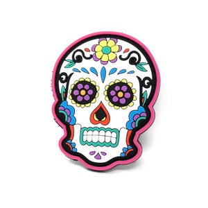 Puede incluir: Un parche de calavera de azúcar colorido con detalles en rosa, blanco, azul, amarillo, verde y morado. La calavera tiene una nariz en forma de corazón y una sonrisa  dentada.