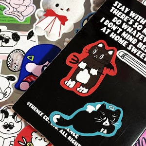 Könnte beinhalten: Ein schwarzer Aufkleber mit einer weißen Katze, die ein rotes Teufelskostüm und einen Heiligenschein trägt. Der Aufkleber befindet sich auf einem schwarzen Hintergrund mit dem Text "Stay with there's so much I don't mind being at home sweet" und "Thence Co., Ltd. All Rights Reserved."
