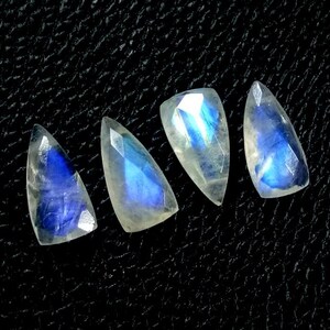 May include: Four teardrop-shaped moonstone gemstones with a faceted cut. The stones have a translucent appearance with a blue and white iridescent glow. The gemstones are arranged on a black textured surface.