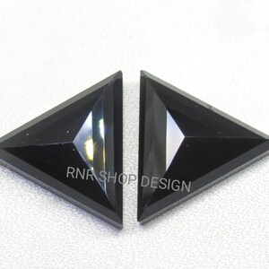 May include: Two black triangular shaped gemstones with a faceted cut. The gemstones are on a white surface. RNR SHOP DESIGN is printed on the surface.