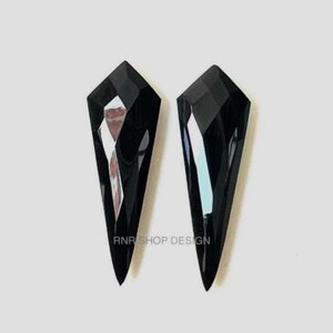 May include: Two black, faceted crystal pendants with an elongated, geometric shape. The crystals have a glossy finish and reflect light, creating a sparkling effect. The pendants are displayed against a plain white background. The text "RNR SHOP DESIGN" is visible.