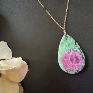 Könnte beinhalten: Eine teardrop-förmige Halskette mit einem grünen und rosafarbenen Edelstein-Anhänger und einer goldenen Kette. Der Edelstein hat ein rosafarbenes blumenartiges Muster in der Mitte.