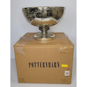 May include: A silver-toned decorative bowl with a pedestal base, sitting atop a cardboard box. The bowl has a wide, rounded shape with a distressed finish. The box is labeled "POTTERYBARN" in black text.
