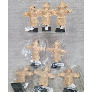 May include: A collection of eight scarecrow figurines crafted from tan burlap fabric. Three figurines stand upright on black metal bases, while five are individually wrapped in clear plastic packaging, each featuring a pointed hat and tied waist.