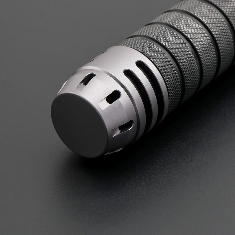 May include: Close-up of a metallic gray cylindrical object with a textured grip and a rounded end. The object has a series of vertical slots and circular openings near the end, and a diamond pattern on the grip.