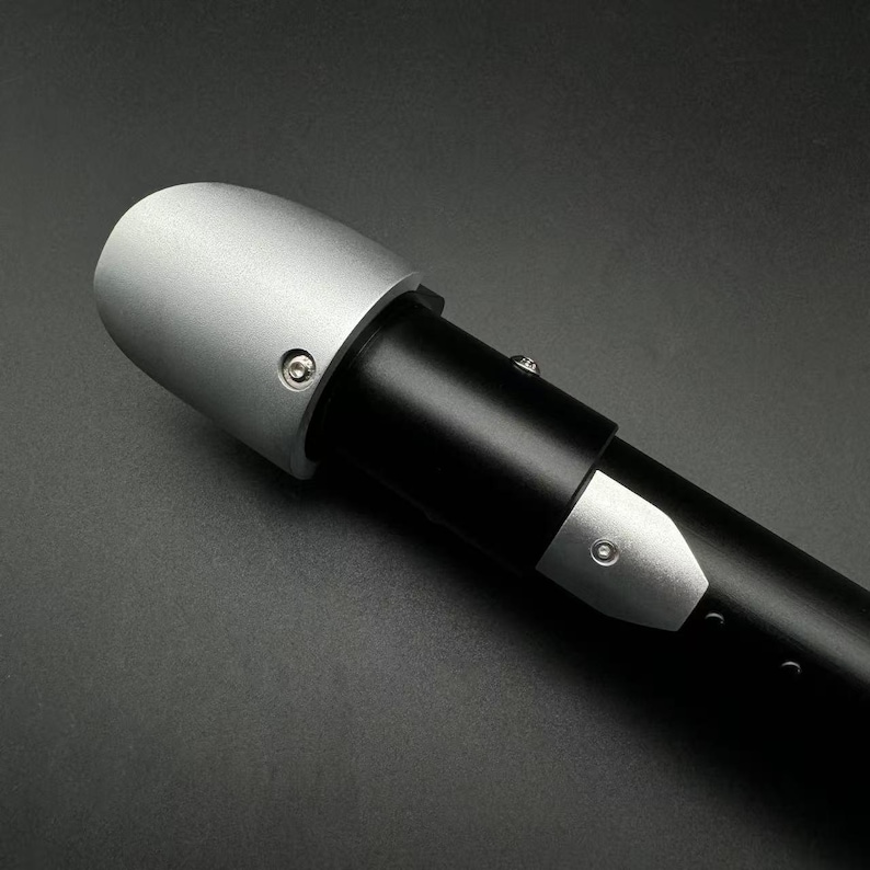 May include: A close-up of a black and silver lightsaber hilt. The hilt is mainly black with silver detailing, including a silver cap and a silver plate. The design is sleek and modern, with visible screws.