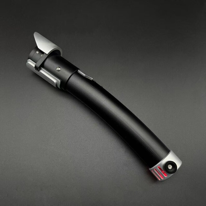 May include: A black lightsaber with silver detailing. The hilt is black with a curved design, and the emitter is silver. The base of the hilt has a red light. The lightsaber is a prop or toy.