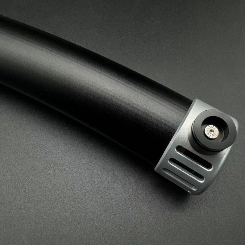 May include: A close-up shot of a black cylindrical object with a silver-coloured end piece. The end piece features a circular design with a screw and vertical slots. The object is set against a dark grey background.