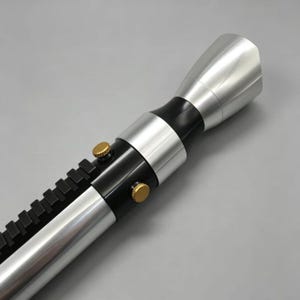 May include: A silver and black lightsaber prop with gold-colored buttons and a black rail. The lightsaber has a metallic finish and a flared emitter.