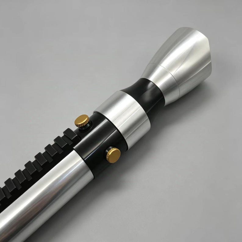 May include: A silver and black lightsaber prop with gold-colored buttons and a black rail. The lightsaber has a metallic finish and a flared emitter.
