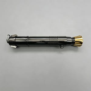 Op de afbeelding: Een zwart en goudkleurig lichtzwaard rekwisiet met een metallic afwerking. Het handvat is zwart met zilveren accenten en een gouden emitter. Het lichtzwaard heeft een ring en een knop aan de zijkant. Het algehele ontwerp is strak en futuristisch.