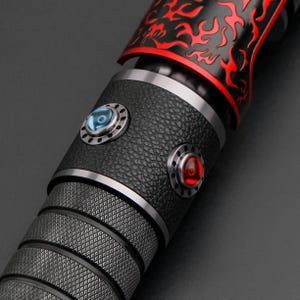 May include: Close-up of a black and red lightsaber hilt. The hilt features a textured grip, a black leather-like section with two circular buttons, one blue and one red. The top section is black with red flame-like designs.