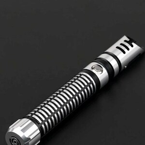 Pu&ograve; includere: Un'elica di spada laser argento e nera con un design dettagliato. L'impugnatura presenta un motivo a coste e un pulsante, mentre la parte superiore ha un design a forma di fessura. La spada laser &egrave; su uno sfondo nero.