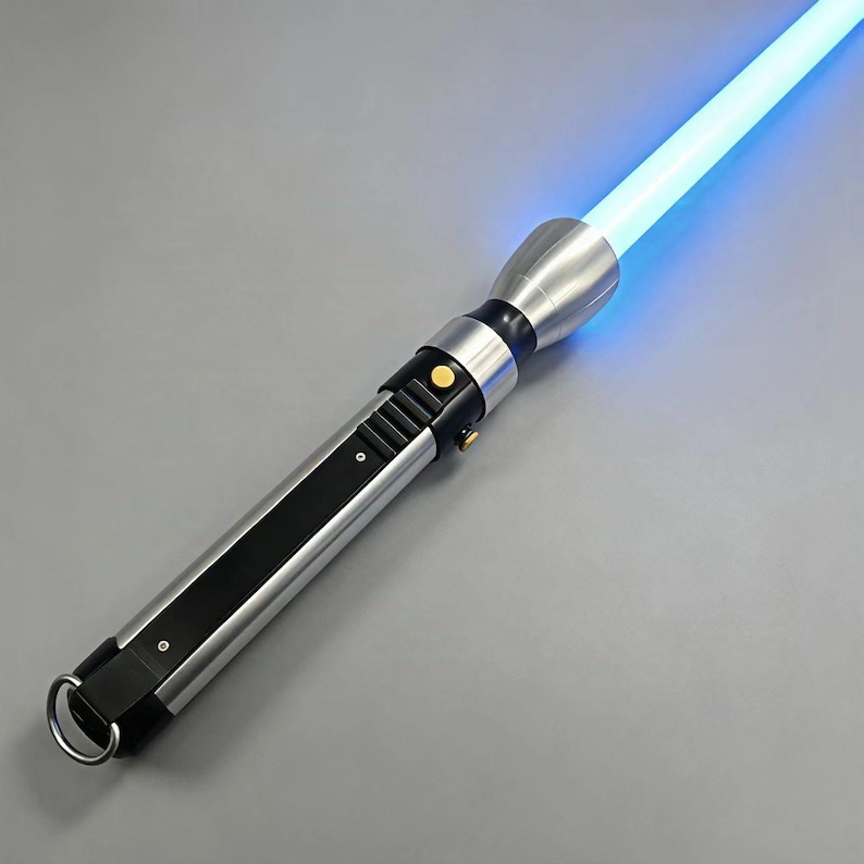 May include: A silver and black lightsaber with a bright blue blade. The hilt features a ring at the base, a black stripe, and several buttons and switches. The glowing blade extends from the top of the hilt.