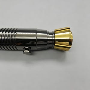 Op de afbeelding: Een metalen lichtzwaardgreep met een zwart-zilveren behuizing en een goudkleurige emitter. De greep heeft een geribbeld ontwerp en een kleine knop. Het lichtzwaard is een rekwisiet of verzamelobject.