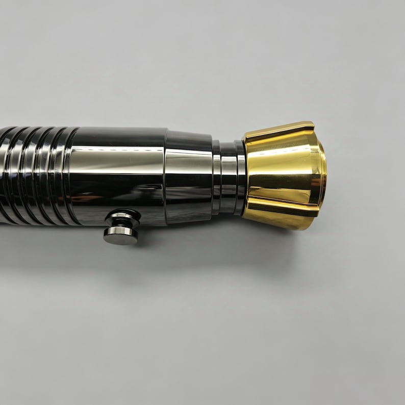Op de afbeelding: Een metalen lichtzwaardgreep met een zwart-zilveren behuizing en een goudkleurige emitter. De greep heeft een geribbeld ontwerp en een kleine knop. Het lichtzwaard is een rekwisiet of verzamelobject.