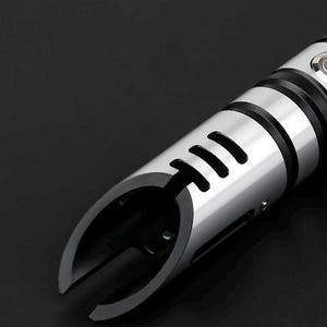 Pu&ograve; includere: Primo piano di un'elsa di spada laser argento e nera. L'elsa ha una finitura metallica con dettagli neri e un pulsante. L'emettitore presenta un design curvo con tre ritagli rettangolari.