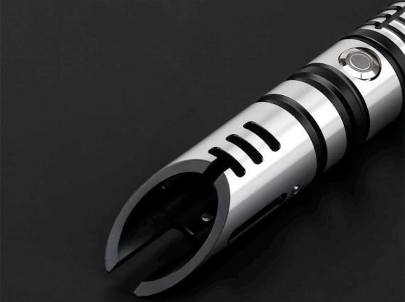 Pu&ograve; includere: Primo piano di un'elsa di spada laser argento e nera. L'elsa ha una finitura metallica con dettagli neri e un pulsante. L'emettitore presenta un design curvo con tre ritagli rettangolari.