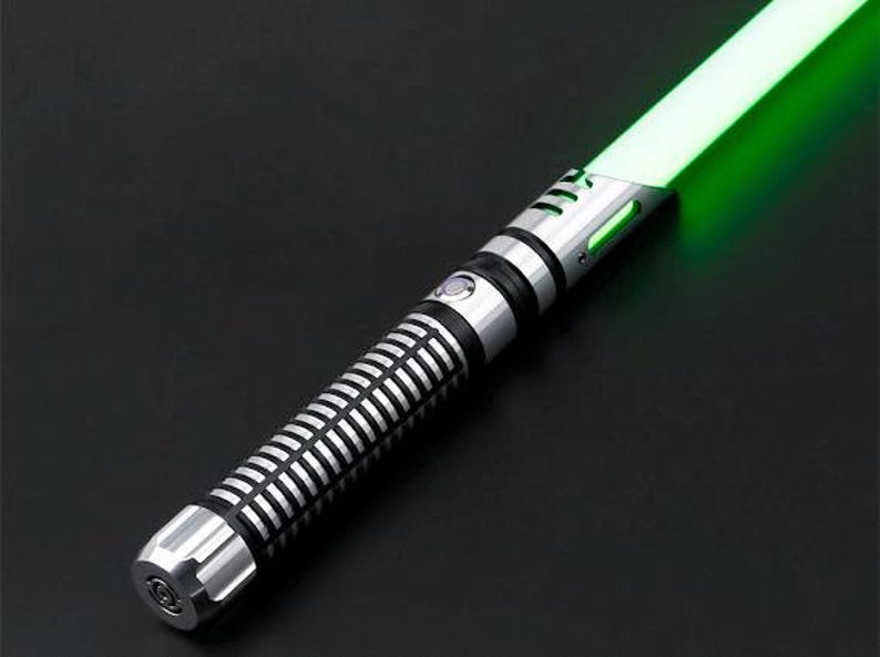 Pu&ograve; includere: Una spada laser argento e nera con una lama verde brillante. L'elsa presenta un design a coste e un pulsante circolare. La lama emette un bagliore verde vibrante su uno sfondo nero.