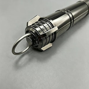 Op de afbeelding: Een zilverkleurig cilindrisch object met een ring eraan bevestigd. Het object heeft geribbelde secties en een complex ontwerp met rechthoekige en cirkelvormige elementen. De algehele esthetiek is industrieel en futuristisch.