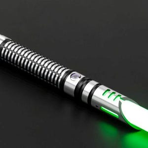 Pu&ograve; includere: Una spada laser argentata e nera con una lama verde brillante. L'elsa ha un design a coste e un pulsante. La lama si illumina intensamente, suggerendo che sia attivata. La spada laser &egrave; ambientata su uno sfondo scuro.